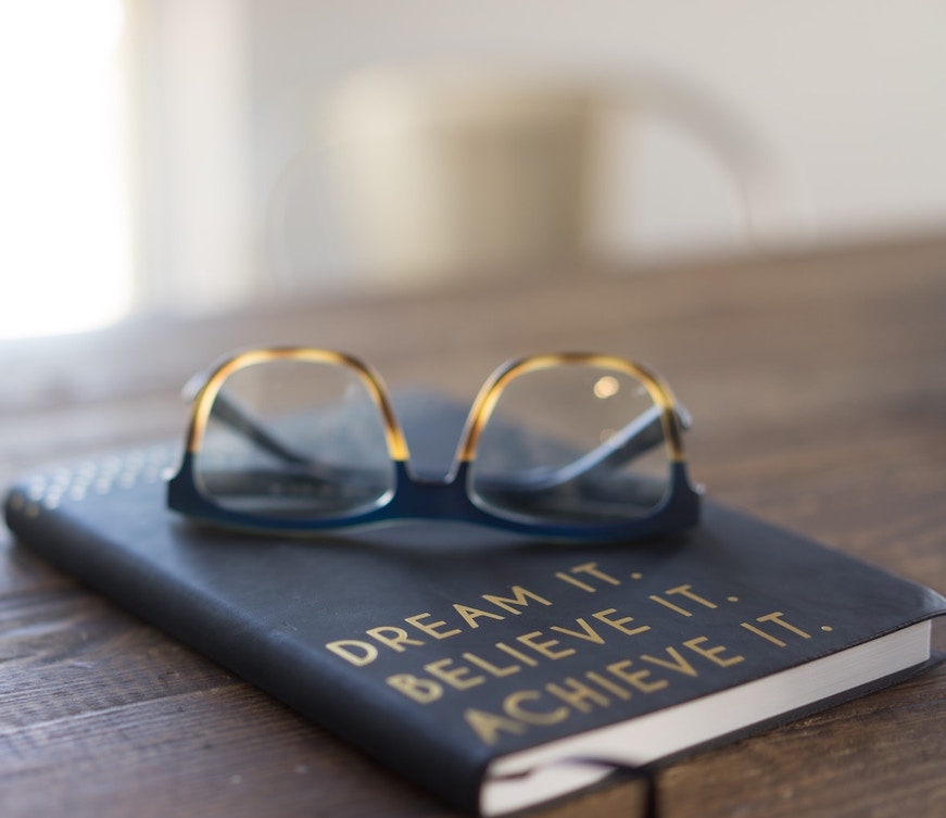 Notebook reading Dream It, Believe It, Achieve It, with a pair of glasses on top.