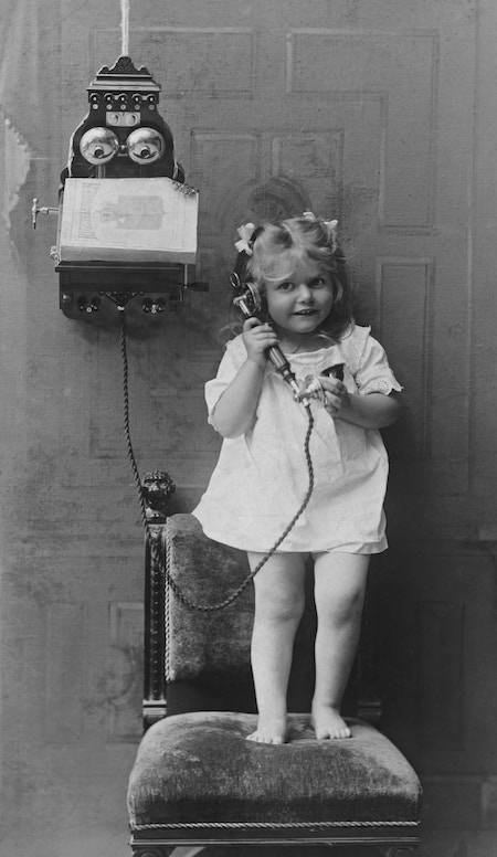 Young girl standing on a chair with the receiver of an old fashion, wall mounted phone.
