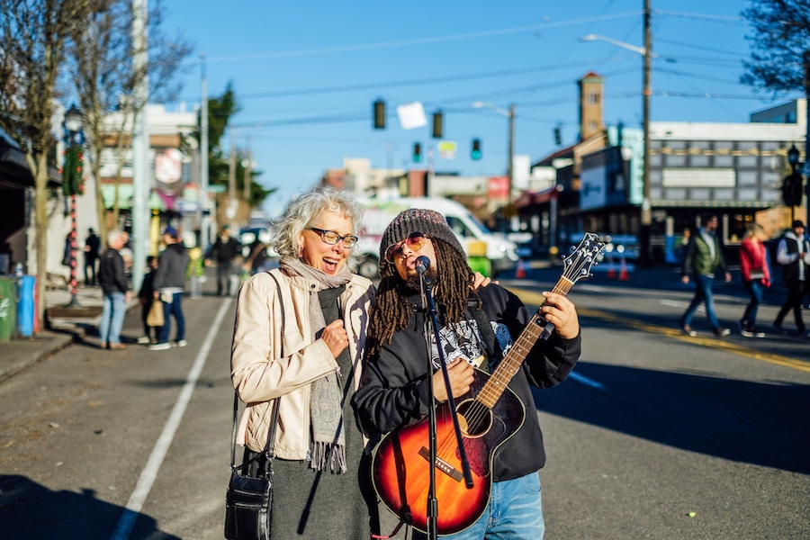 Older woman and younger man in dreadlocks with a guitar singing together in the streets.