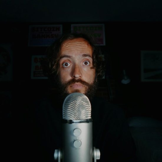 Wide eyed man, shrouded by darkness, in front of a microphone.